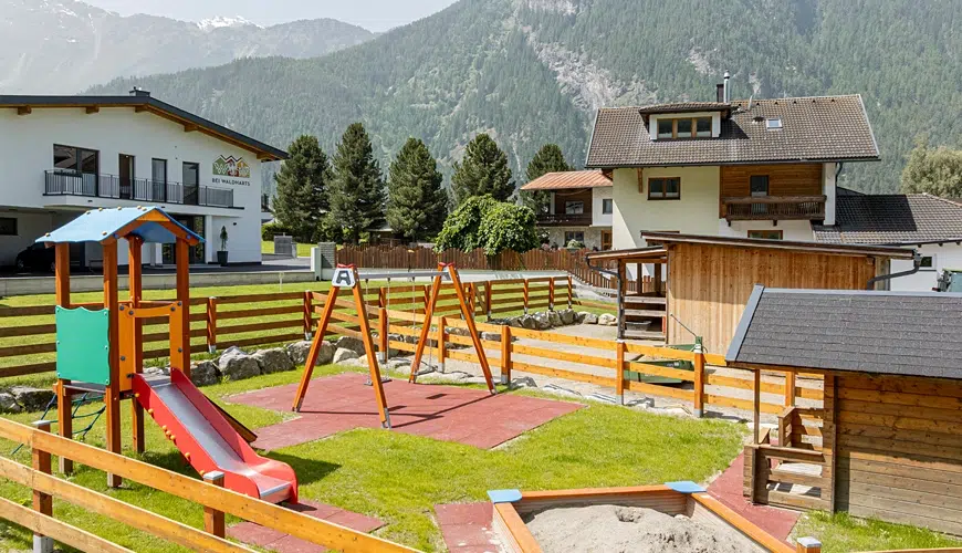 Ein eingezäunter Spielplatz mit Schaukeln, Rutsche, Sandkasten und nahe gelegenen Häusern in einem Berggebiet.