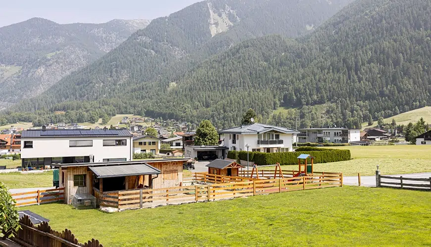 Ein eingezäunter Hof mit Tiergehegen, Häusern und Bergen im Hintergrund an einem sonnigen Tag.
