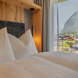 Cozy bed with white linens next to a window overlooking mountains and village houses.