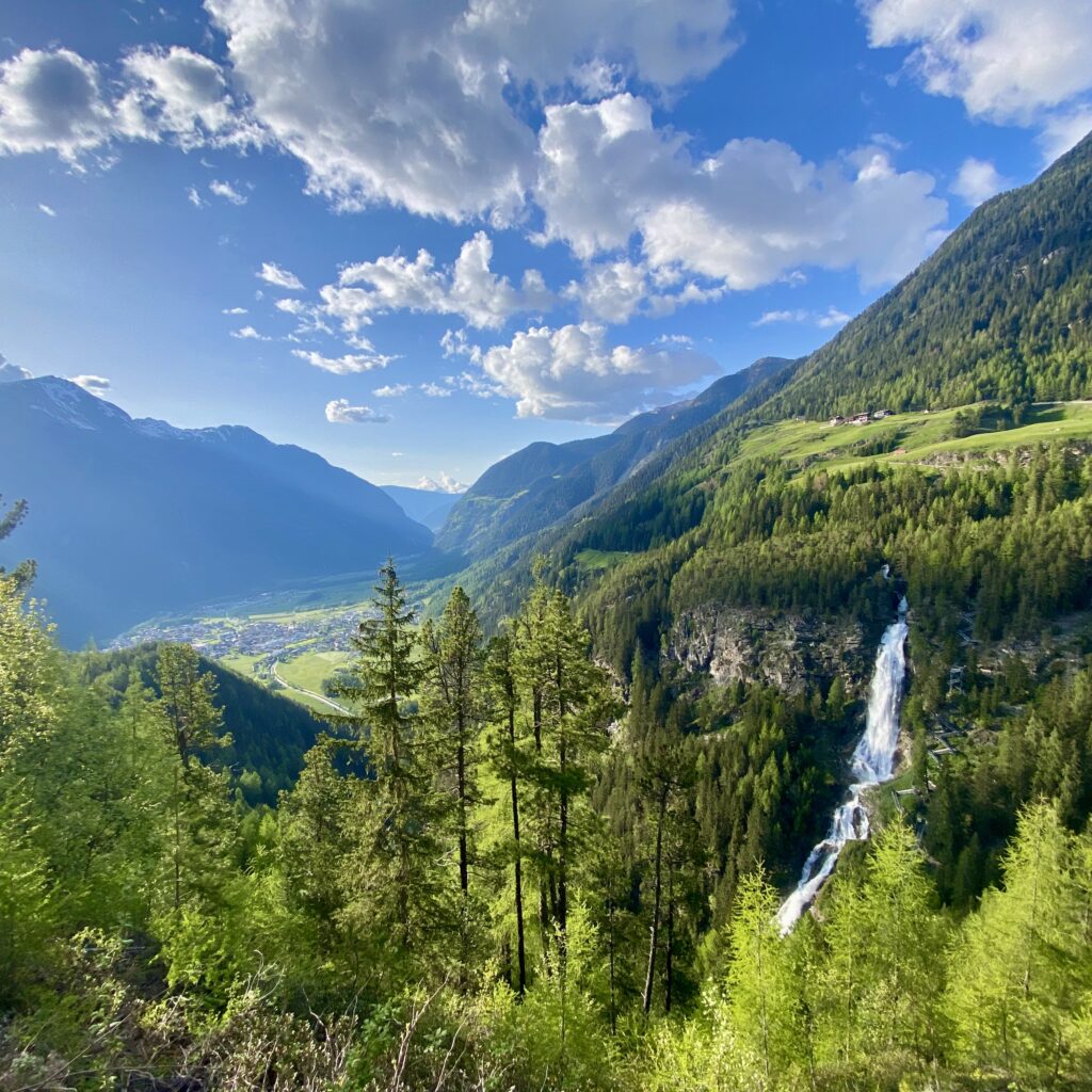 Ein üppig grünes Tal mit hohen Bäumen, einem Wasserfall und den Bergen des Ötztals.